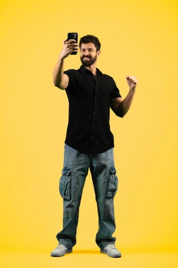 استخدام الهاتف الجوال, رفع قبضة اليد للأعلى بإيماءات الفوز، إجراء بث مباشر عبر وسائل التواصل الاجتماعي, بوتريه لشاب سعودي يرتدي الزي الكاجوال يقوم بالتقاط صور تذكارية، رجل عربي اماراتي خليجي يقوم بالرد على مكالمة فيديو هاتفية، صورة الجسم بالكامل، خلفية صفراء