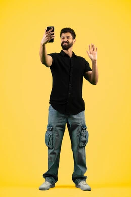 إلقاء التحية والسلام بإيماءات البشاشة،استخدام الهاتف الجوال, إجراء بث مباشر عبر وسائل التواصل الاجتماعي, بوتريه لشاب سعودي يرتدي الزي الكاجوال يقوم بالتقاط صور تذكارية، رجل عربي اماراتي خليجي يقوم بالرد على مكالمة فيديو هاتفية، صورة الجسم بالكامل، خلفية صفراء