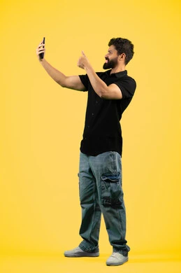 رفع إصبع الابهام بايماءات الاعجاب،استخدام الهاتف الجوال, إجراء بث مباشر عبر وسائل التواصل الاجتماعي, بوتريه لشاب سعودي يرتدي الزي الكاجوال يقوم بالتقاط صور تذكارية، رجل عربي اماراتي خليجي يقوم بالرد على مكالمة فيديو هاتفية، صورة الجسم بالكامل، خلفية صفراء