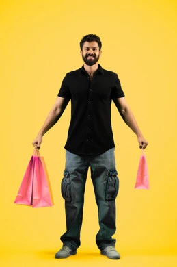 الاستفادة من مواسم الحسومات والتصفيات، المشتريات التجارية والهدايا, شاب سعودي ينظر إلى الكاميرا بتعابير السرور، التبضع وأخذ الاحتياجات اللازمة, بورتريه لرجل عربي خليجي اماراتي يرتدي الزي الكاجوال و يمسك بيده أكياس ورقية، صورة الجسم بالكامل، خلفية صفراء