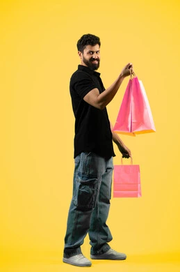 شراء الاحتياجات اللازمة, الاستفادة من مواسم الحسومات والتصفيات، المشتريات التجارية والهدايا, شاب سعودي ينظر إلى الكاميرا بتعابير السرور، بورتريه من الجانب لرجل عربي خليجي اماراتي يرتدي الزي الكاجوال و يمسك بيده أكياس ورقية، صورة الجسم بالكامل، خلفية صفراء
