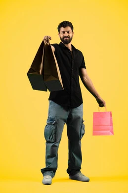 Business purchases and gifts, a young Saudi man looking at the camera with a happy expression, shopping and taking the necessary supplies, seasons of offers and discounts, taking advantage of discounts and clearance sales, an Arab Gulf Emirati man wearing casual clothes and holding paper bags in his hand, full body image, yellow background