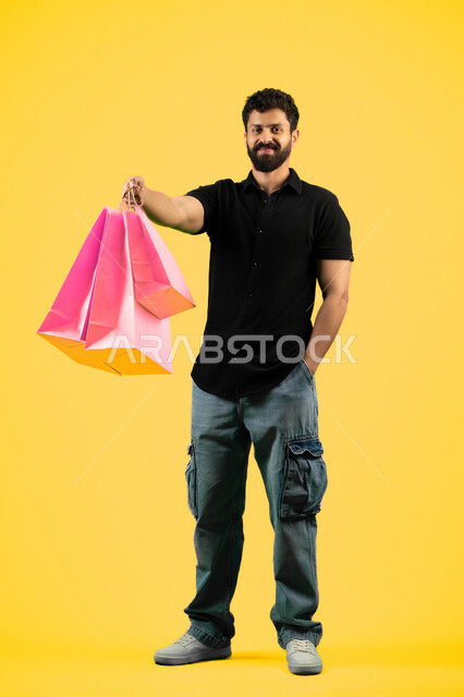 التبضع وأخذ الاحتياجات اللازمة, الاستفادة من مواسم الحسومات والتصفيات، المشتريات التجارية والهدايا, شاب سعودي ينظر إلى الكاميرا بتعابير السرور، بورتريه لرجل عربي خليجي اماراتي يرتدي الزي الكاجوال و يمسك بيده أكياس ورقية، صورة الجسم بالكامل، خلفية صفراء