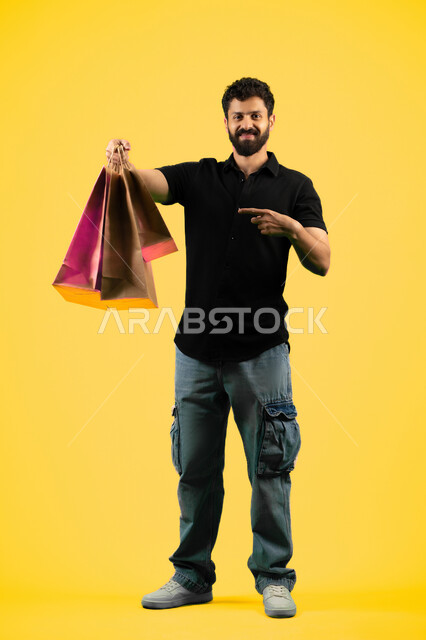 Pointing index finger at commercial purchases and gifts, taking advantage of discount seasons, a young Saudi man looking at the camera with an expression of pleasure, side portrait of an Arab Gulf Emirati man wearing casual clothes and holding paper bags, full body photo, yellow background