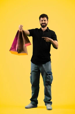 الاشارة بإصبع السبابة إلى المشتريات التجارية والهدايا, الاستفادة من مواسم الحسومات، شاب سعودي ينظر إلى الكاميرا بتعابير السرور، بورتريه من الجانب لرجل عربي خليجي اماراتي يرتدي الزي الكاجوال و يمسك بيده أكياس ورقية، صورة الجسم بالكامل، خلفية صفراء