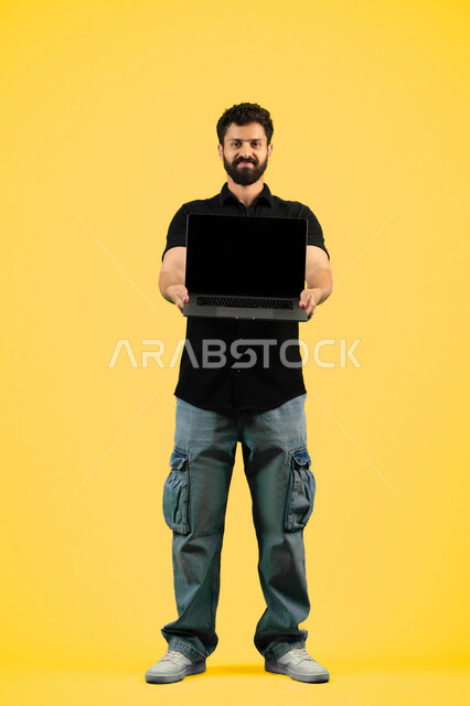 استخدام الأجهزة الحديثة في انجاز المهام, بورتريه لرجل عربي خليجي اماراتي يرتدي الزي الكاجوال يحمل الحاسوب المحمول، شاب سعودي يعرض شاشة سوداء فارغة عبر اللابتوب، الجسم بالطول الكامل، خلفيه صفراء