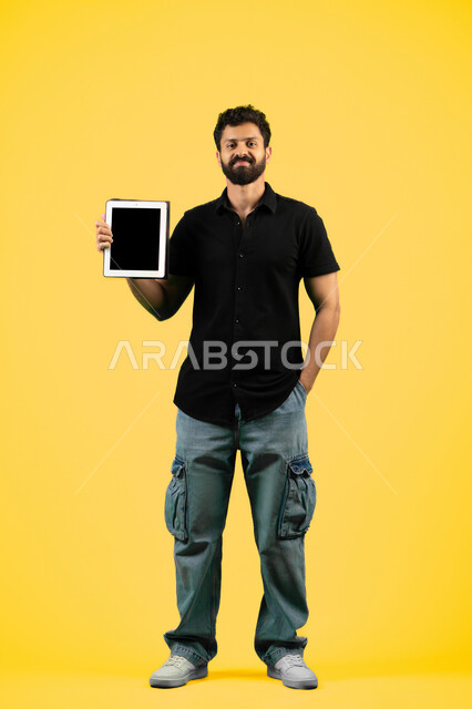 Looking at the camera with happy gestures, using modern and advanced technology, a Saudi young man showing a blank black screen via tablet, portrait of an Arab Gulf Emirati man wearing casual clothes holding a tablet in his hand, full body length, yellow background