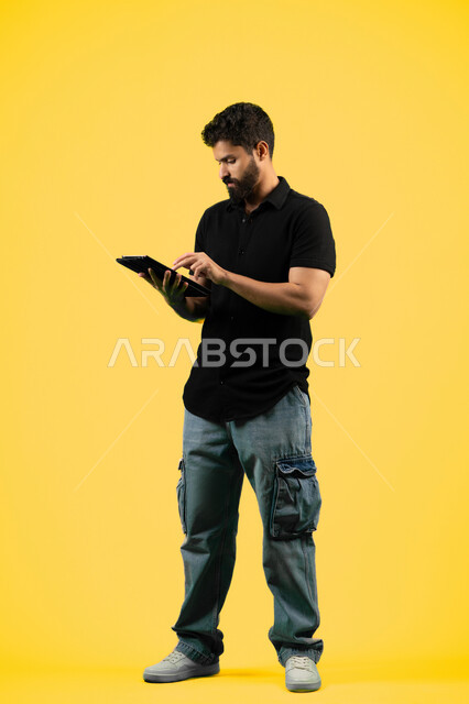 استخدام الجهاز اللوحي في إنجاز مهام العمل، شاب سعودي يتصفح تطبيقات التواصل الاجتماعي، استعمال التكنولوجيا الحديثة والمتطورة، بورتريه لرجل عربي خليجي اماراتي يرتدي الزي الكاجوال يستخدم جهاز التابلت بإيماءات التركيز، طول الجسم بالكامل، خلفية صفراء