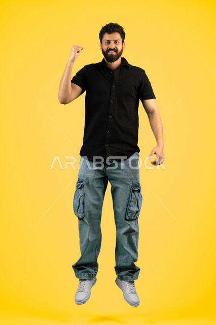 Victory and triumph of the favorite team, mastery of boxing arts, increased muscle endurance, portrait of a smiling young Arab Gulf Emirati man in casual clothes raising his fists with gestures of strength and victory, a Saudi man looking at the camera with an expression of self-confidence, full body, yellow background