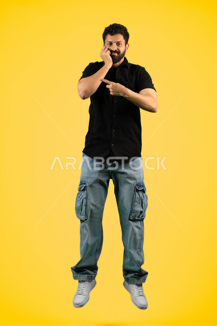 The concept of elegance and interest in outward appearance, gestures of boredom and disappointment, portrait of a smiling young Arab Gulf Emirati man wearing casual clothes standing straight and looking at the camera, a Saudi man putting his hand on his face and pointing with his index finger at something, full body, yellow background