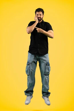 The concept of elegance and interest in outward appearance, gestures of boredom and disappointment, portrait of a smiling young Arab Gulf Emirati man wearing casual clothes standing straight and looking at the camera, a Saudi man putting his hand on his face and pointing with his index finger at something, full body, yellow background