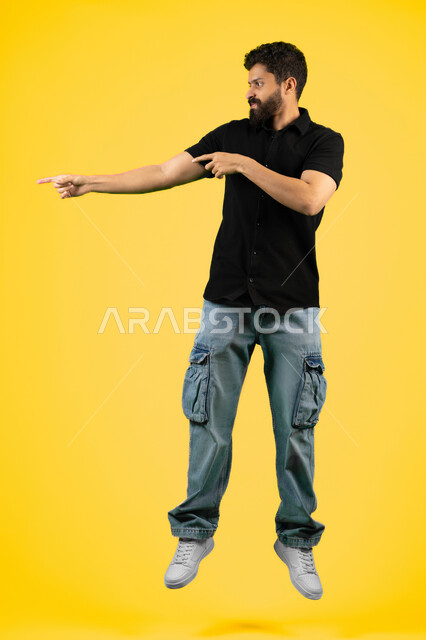 Pointing to something specific, elegance and interest in outward appearance, portrait of a smiling young Arab Gulf Emirati man wearing casual clothes standing straight, Saudi man pointing with his index finger at something, full body, yellow background
