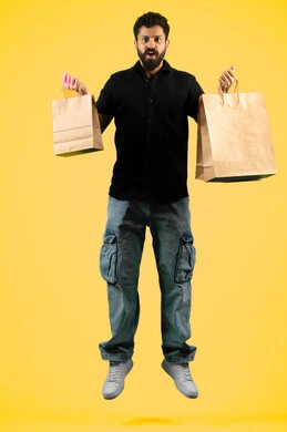 الاستفادة من مواسم الحسومات والتصفيات، المشتريات التجارية والهدايا, شاب سعودي ينظر إلى الكاميرا بتعابير السرور، التبضع وأخذ الاحتياجات اللازمة, بورتريه لرجل عربي خليجي اماراتي يرتدي الزي الكاجوال و يمسك بيده أكياس ورقية، صورة الجسم بالكامل، خلفية صفراء