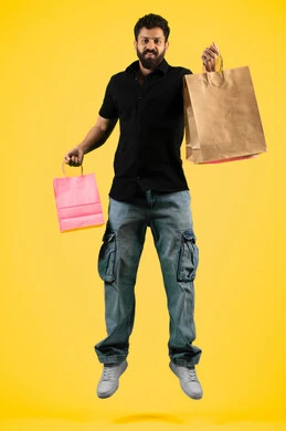 Business purchases and gifts, a young Saudi man looking at the camera with a happy expression, shopping and taking the necessary supplies, seasons of offers and discounts, taking advantage of discounts and clearance sales, an Arab Gulf Emirati man wearing casual clothes and holding paper bags in his hand, full body image, yellow background