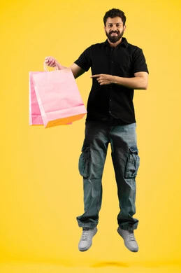 Pointing index finger at commercial purchases and gifts, taking advantage of discount seasons, a young Saudi man looking at the camera with an expression of pleasure, side portrait of an Arab Gulf Emirati man wearing casual clothes and holding paper bags, full body photo, yellow background