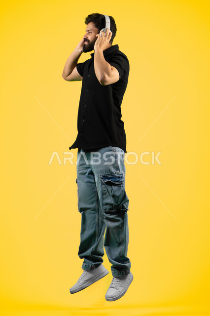 Artistic singing talents, an Arab Gulf Emirati man wearing headphones, gestures of happiness and integration with the song, a portrait of a Saudi young man wearing casual clothes listening to music, a recording of a singing audio clip, a full body image, a yellow background