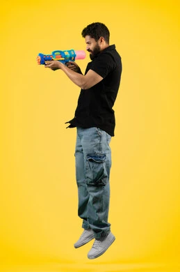 Having fun with water games in summer, concept of entertainment and amusement, side portrait of a young Arab Gulf Emirati man wearing casual clothes holding a plastic water gun playing with gestures of pleasure and happiness, full body, braided background