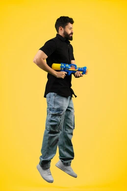 قضاء وقت ممتع بالألعاب المائية في فصل الصيف، مفهوم الترفيه عن النفس والتسلية، بورتريه من الجانب لشاب عربي خليجي اماراتي يرتدي الزي الكاجوال يحمل مسدس مائي بلاستيكي يقوم باللعب بإيماءات السرور والسعادة، الجسم بالطول الكامل، خلفية ضفراء