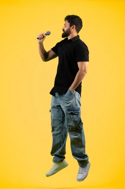 الغناء بحماس وصوت عالي، الفن المسرحي الغنائي، تقديم العروضات المسرحية، رجل اماراتي يقوم بممارسة هواية الغناء للتسلية والترفيه، بورتريه لشاب عربي سعودي خليجي يرتدي الزي الكاجوال يحمل بيده الميكروفون، مواهب فنية في عالم الموسيقى، طول الجسم بالكامل، خلفية صفراء