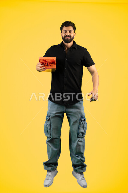Expressing love, appreciation and respect, giving gifts on holidays and special occasions, concept of thanks and gratitude, Saudi man looking at the camera with gestures of joy and happiness, portrait of a young Arab Gulf Emirati man wearing casual clothes holding a gift box, full body, yellow background