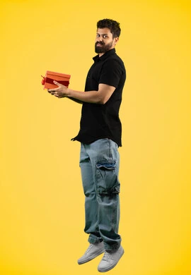 مفهوم الشكر والامتنان، التعبير عن الحب والتقدير، تقديم الهدايا بالاعياد والمناسبات الخاصة، رجل سعودي ينظر إلى الكاميرا بايماءات السرور، بورتريه من الجانب  لشاب عربي خليجي اماراتي يرتدي ازي الكاجوال يحمل بيديه صندوق هدية، الجسم بالطول الكامل، خلفية صفراء