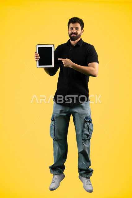 Pointing index finger at tablet, using modern and advanced technology, Saudi young man showing blank black screen on tablet, portrait of an Arab Gulf Emirati man in casual wear looking at camera with happy gestures, full body length, yellow background