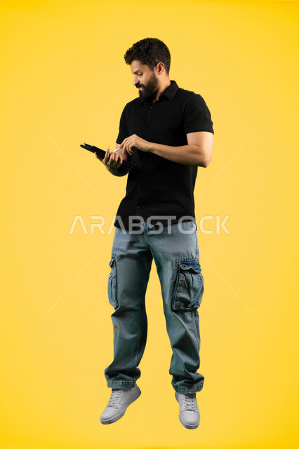 Using modern and advanced technology, using a tablet to complete work tasks, a Saudi young man browsing social media applications, a portrait of an Arab Gulf Emirati man wearing casual clothes writing with a pen on a tablet with focus gestures, full body length, yellow background