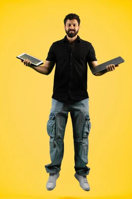 استخدام الأجهزة المحمولة في إنجاز مهام العمل، شاب سعودي ينظر إلى الكاميرا بإيماءات الاعجاب بالتكنولوجيا الحديثة والمتطورة، بورتريه لرجل عربي خليجي اماراتي يرتدي الزي الكاجوال يحمل جهاز اللابتوب والتابليت، طول الجسم بالكامل، خلفية صفراء