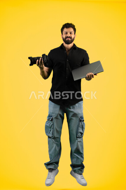 Mastering the art of photography, a young Saudi man looking at the camera with gestures of admiration for modern and advanced technology, portrait of an Arab Gulf Emirati man in casual wear holding a laptop and a high-resolution digital camera, full body length, yellow background