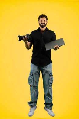 Mastering the art of photography, a young Saudi man looking at the camera with gestures of admiration for modern and advanced technology, portrait of an Arab Gulf Emirati man in casual wear holding a laptop and a high-resolution digital camera, full body length, yellow background