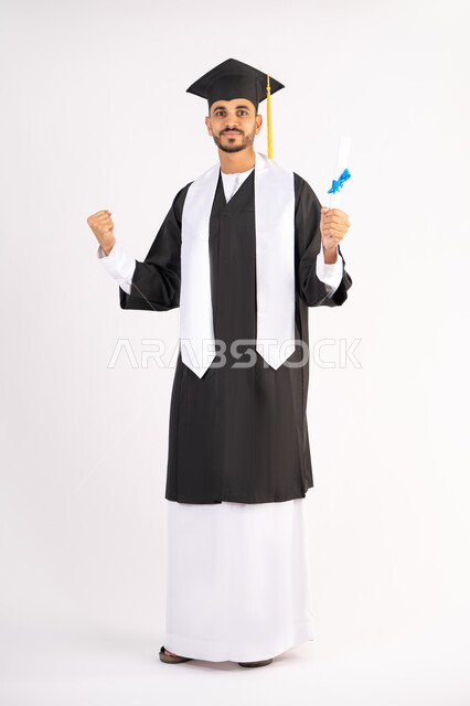 رفع قبضة اليد بتعابير الانجاز، الفخر بالوصول إلى الأحلام والنجاح، النظر إلى الكاميرا بإيماءات السعادة والفرح، الاحتفال بإنهاء المرحلة الجامعية، بورتريه لشب خريج عربي خليجي اماراتي يرتدي القبعة وعباءة التخرج يحمل بيده الشهادة، الجسم بالطول الكامل، خلفية بيضاء