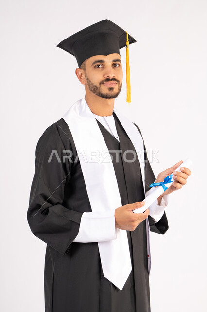 Celebrating the completion of university, expressions of joy and happiness for success, taking selfies in graduation clothes, portrait of a smiling young Arab Gulf Emirati graduate wearing a graduation cap and gown, holding a certificate, looking at the camera with gestures of joy, white background