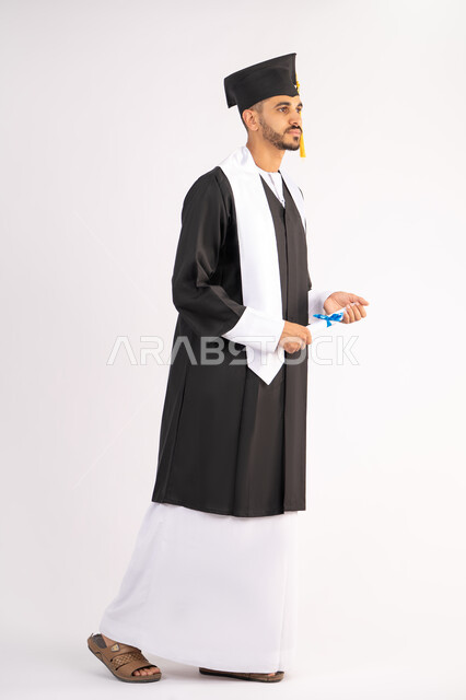 Achieving the dream of success, expressions of happiness and pleasure, university occasion and celebration, celebration of completing university studies, concept of diligence and excellence, portrait of a smiling young Arab Gulf Emirati graduate wearing a graduation gown holding a certificate in his hand, full body, white background