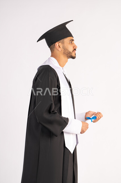 Holding a certificate with expressions of joy and happiness, feeling proud and self-confident, celebrating success and completing university studies, the concept of excellence and diligence, a portrait of an Arab Gulf Emirati graduate wearing a graduation cap and gown, standing straight with gestures of happiness, white background