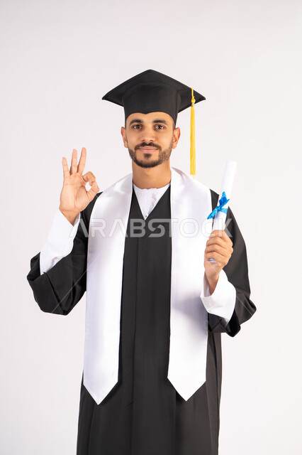 علامة الجودة والامتياز والتميز، الاحتفال بالوصول إلى الأحلام، الاحتفال بإنهاء المرحلة الجامعية، بورتريه لشاب خريج عربي خليجي اماراتي يرتدي القبعة وعباءة التخرج يحمل الشهادة بيده ينظر إلى الكاميرا بإيماءات السعادة، خلفية بيضاء