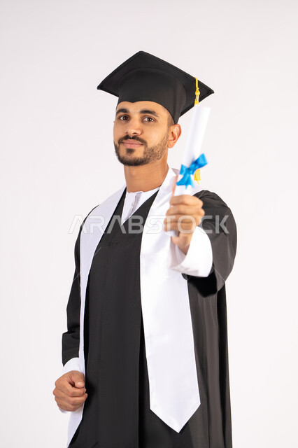 تعابير السعادة والسرور، مناسبة واحتفال جامعي، الاحتفال بإنهاء الدراسة الجامعية، مفهوم الاجتهاد والتفوق، بورتريه لشاب خريج عربي خليجي اماراتي مبتسم يرتدي عباءة التخرج يمسك بيده الشهادة، الوصول لحلم النجاح،  خلفية بيضاء