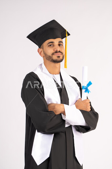 التقاط صور شخصية بثياب التخرج، الاحتفال بإنهاء المرحلة الجامعية، بورتريه لشاب خريج عربي خليجي اماراتي مبتسم يرتدي القبعة وعباءة التخرج يقف بأيدي متقاطعة ينظر الى الكاميرا بتعابير الثقة بالنفس، مفهوم التفوق والنجاح والاجتهاد، خلفية بيضاء
