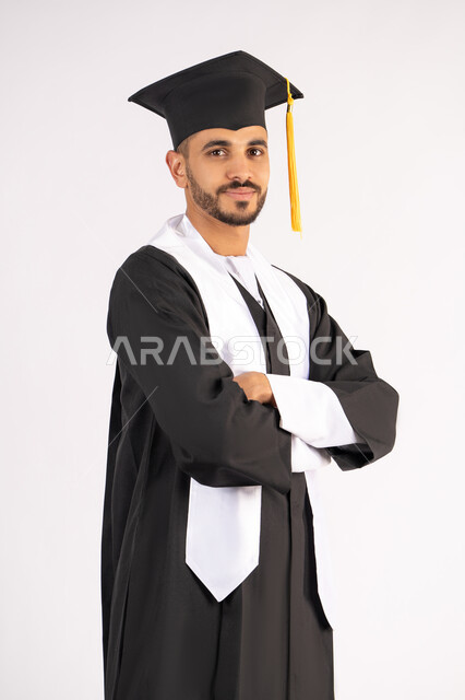 التقاط صور شخصية بثياب التخرج، الاحتفال بإنهاء المرحلة الجامعية، بورتريه لشاب خريج عربي خليجي اماراتي مبتسم يرتدي القبعة وعباءة التخرج يقف بأيدي متقاطعة ينظر الى الكاميرا بتعابير الثقة بالنفس، مفهوم التفوق والنجاح والاجتهاد، خلفية بيضاء