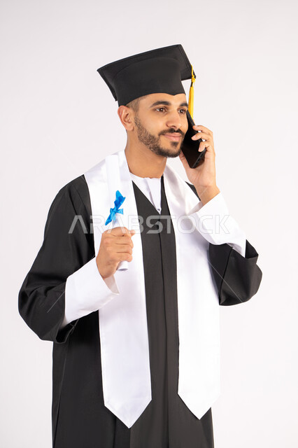 التواصل مع الأهل والأصدقاء، مفهوم الاجتهاد والتفوق، الاحتفال بإنهاء الدراسة الجامعية، استخدام التقنية الحديثة، بورتريه مقرب لشاب خريج عربي خليجي اماراتي يرتدي عباءه وقبعة التخرج يقوم بإجراء مكالمة عبر الهاتف المحمول،  خلفية بيضاء