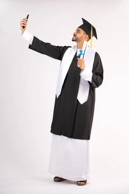 Preserving happy memories, using modern technology, celebrating the completion of university studies, portrait of a young Arab Gulf Emirati graduate wearing an abaya and graduation cap, holding the document in his hand, taking a commemorative selfie with his mobile phone, full body, white background