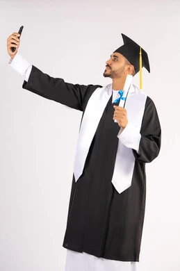 الاحتفاظ بالذكريات السعيدة، استخدام التقنية الحديثة، الاحتفال بإنهاء الدراسة الجامعية، بورتريه لشاب خريج عربي خليجي اماراتي يرتدي عباءه وقبعة التخرج يمسك بيده الوثيقة يقوم بالتقاط صورة سيلفي تذكارية عبر الهاتف المحمول، خلفية بيضاء