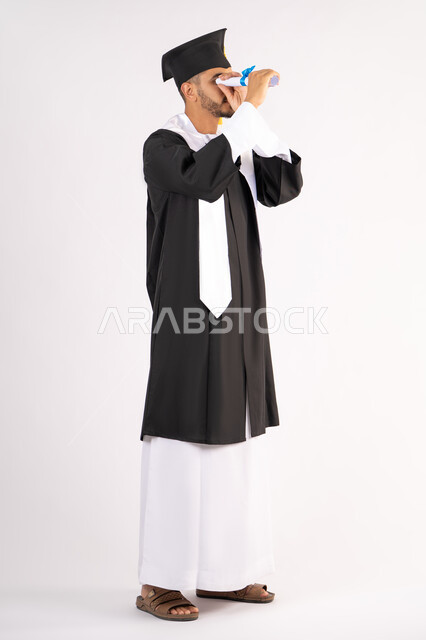 Joy and pride in achieving the dream of success, portrait of a young Saudi Arabian Gulf graduate wearing graduation robes looking through his certificate of excellence, celebrating the end of the university stage, looking forward to the future after graduation, full body, white background