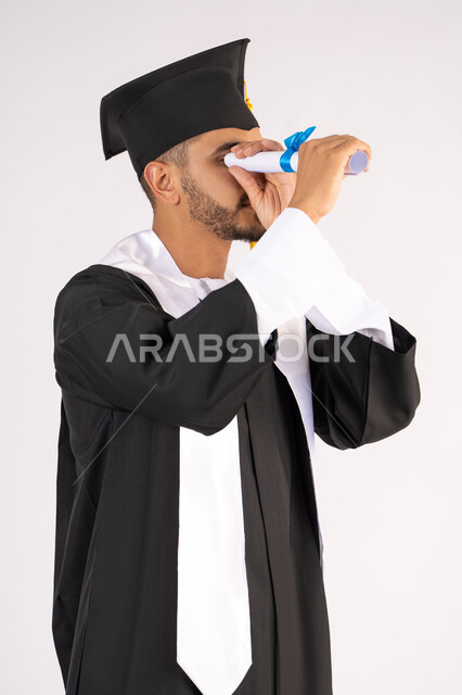 التطلع إلى المستقبل بعد التخرج، الفخر والفرح بالوصول لحلم النجاح، بورتريه لشاب خريج عربي سعودي خليجي يرتدي ثياب التخرج ينظر من خلال شهادة التفوق، الاحتفال بانتهاء المرحلة الجامعية، خلفية بيضاء