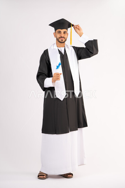 تعابير السعادة والسرور، مناسبة واحتفال جامعي، الاحتفال بإنهاء الدراسة الجامعية، مفهوم الاجتهاد والتفوق، بورتريه لشاب خريج عربي خليجي اماراتي مبتسم يرتدي عباءة التخرج يمسك بيده الشهادة، الوصول لحلم النجاح، الجسم بالطول الكامل، خلفية بيضاء