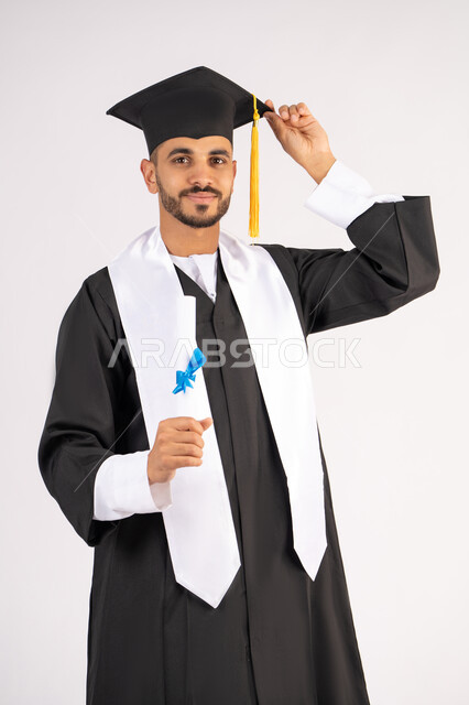 ايماءات السعادة والسرور، مناسبة واحتفال جامعي، الاحتفال بإنهاء الدراسة الجامعية، مفهوم الاجتهاد والتفوق، بورتريه لشاب خريج عربي خليجي اماراتي مبتسم يرتدي عباءة التخرج يمسك بيده الشهادة، الوصول لحلم النجاح، خلفية بيضاء