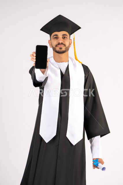 Online education and study programs and applications, portrait of a smiling young Arab Gulf Emirati graduate wearing a cap and graduation gown, holding a certificate with an expression of joy, celebrating the end of the university stage, displaying a blank black screen on a mobile phone, white background