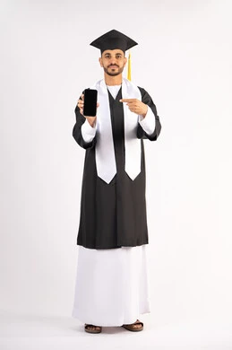 Blank black screen on mobile phone, online education and study applications, portrait of a smiling young Arab Gulf Emirati graduate wearing a cap and graduation gown with a happy expression, celebrating the end of the university stage, full body, white background