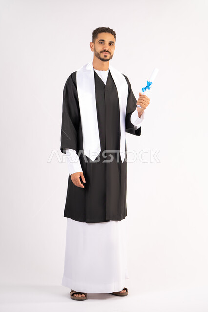 الوصول لحلم النجاح، تعابير السعادة والسرور، مناسبة واحتفال جامعي، الاحتفال بإنهاء الدراسة الجامعية، مفهوم الاجتهاد والتفوق، بورتريه لشاب خريج عربي خليجي اماراتي مبتسم يرتدي عباءة التخرج يمسك بيده الشهادة، الجسم بالطول الكامل، خلفية بيضاء