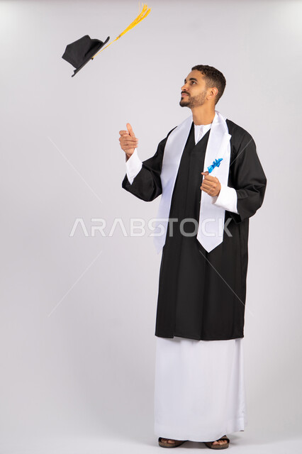 Throwing the hat up with expressions of joy and happiness, quality of education in the Emirates, feeling proud and happy to achieve the dream of graduation, portrait of a young Arab Gulf Emirati graduate wearing an abaya holding a certificate in his hand, celebrating the completion of the university stage, full body, white background