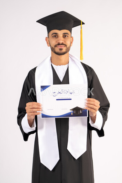 حمل الشهادة بتعابير الفروح والسرور، الشعور بالفخر والثقة بالنفس، الاحتفال بالنجاح وانهاء الدراسة الجامعية، مفهوم التفوق والاجتهاد، بورتريه لخريج عربي خليجي اماراتي يرتدي قبعة وثوب التخرج يقف باستقامة بايماءات السعادة، خلفية بيضاء